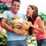 A photo of a man holding a armful of different fruits, while a woman laughs as she adds more to his armful